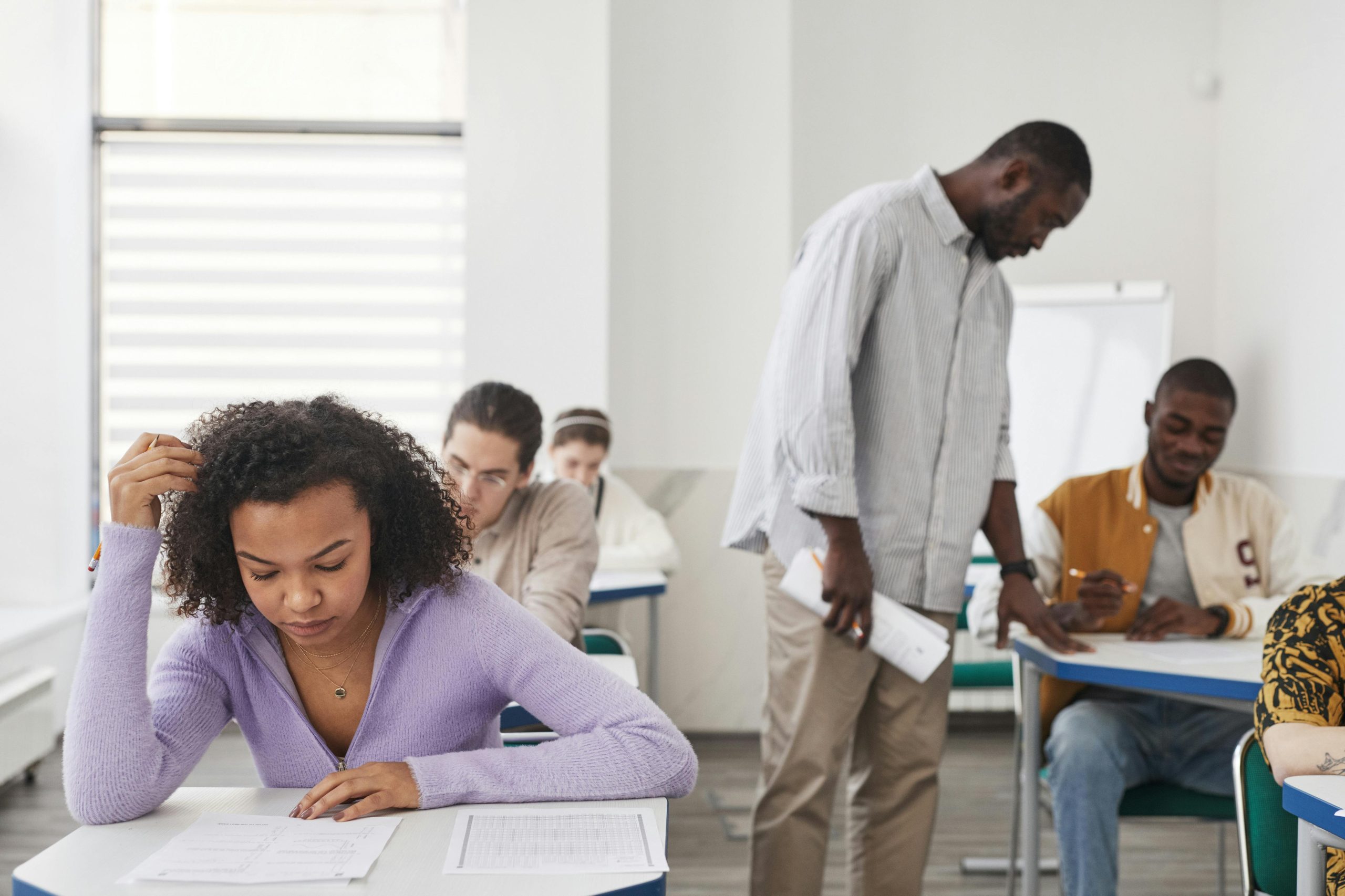 A diverse group of students taking an exam in a modern classroom with a teacher supervising.